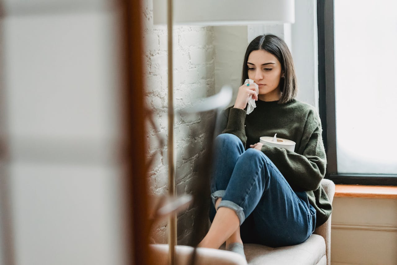 Image of a sad woman holding tissue and cup, sitting curled up on a chair by a window, representing can you forgive someone and still be hurt.