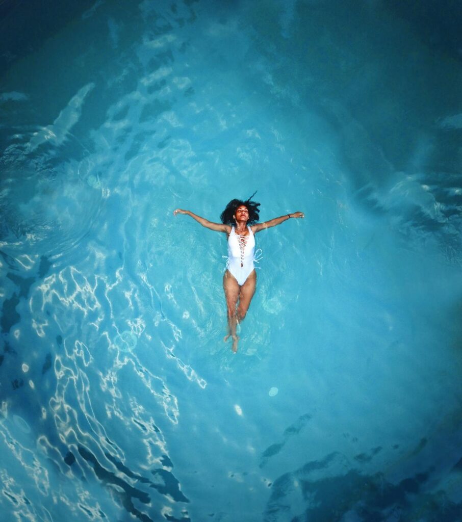 Image of a woman floating peacefully in a bright blue swimming pool wearing a white swimsuit, arms spread wide, showing self care for single moms.