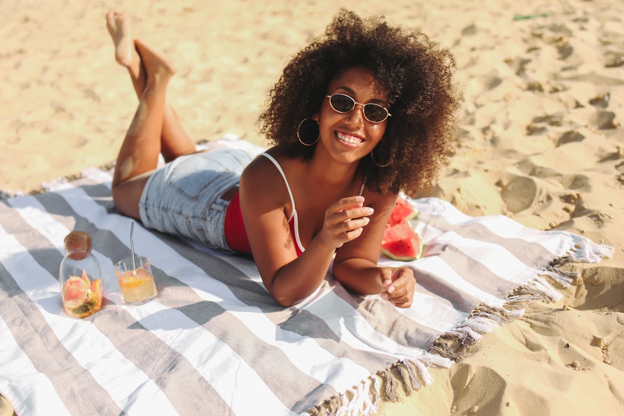 Image of a smiling woman in sunglasses lying on a beach towel with drinks and watermelon slices, representing types of women's shorts.