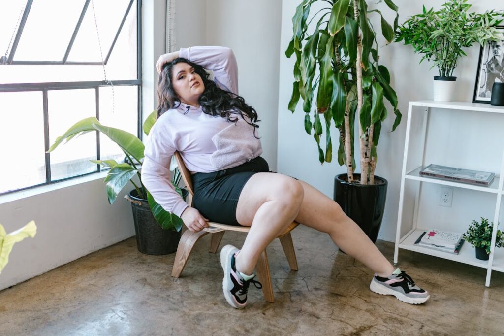 Image of a relaxed woman in casual attire sitting on a wooden chair near potted plants by a large window, showing best short styles for heavy thighs.