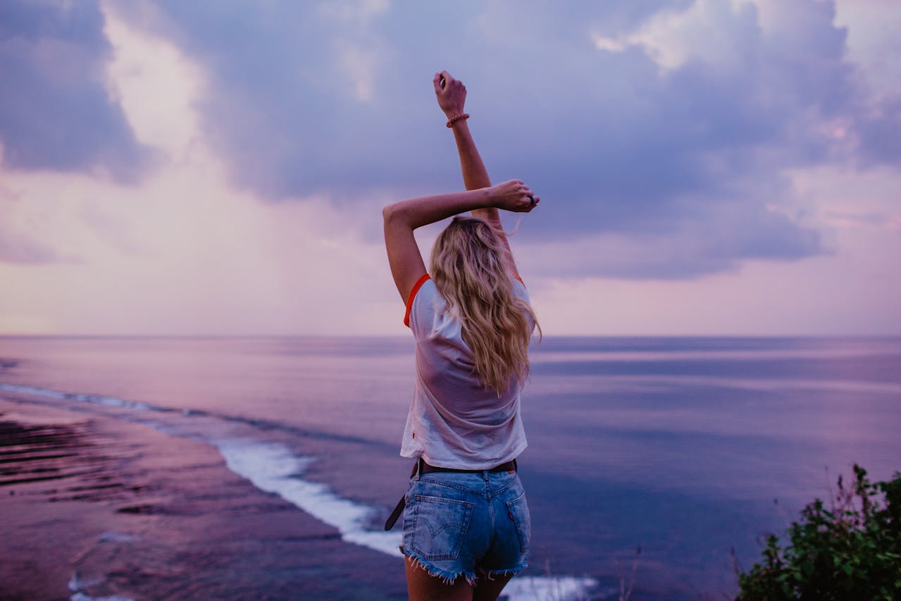 Image of a woman with arms raised facing ocean at sunset, wearing white tee and denim shorts, representing flattering shorts for big thighs.