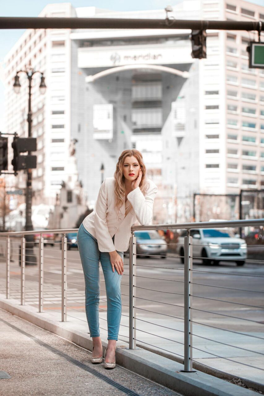 Image of a stylish woman in white blazer and jeans leaning on railing with city buildings in the background, showing clothes to wear in Las Vegas for ladies.