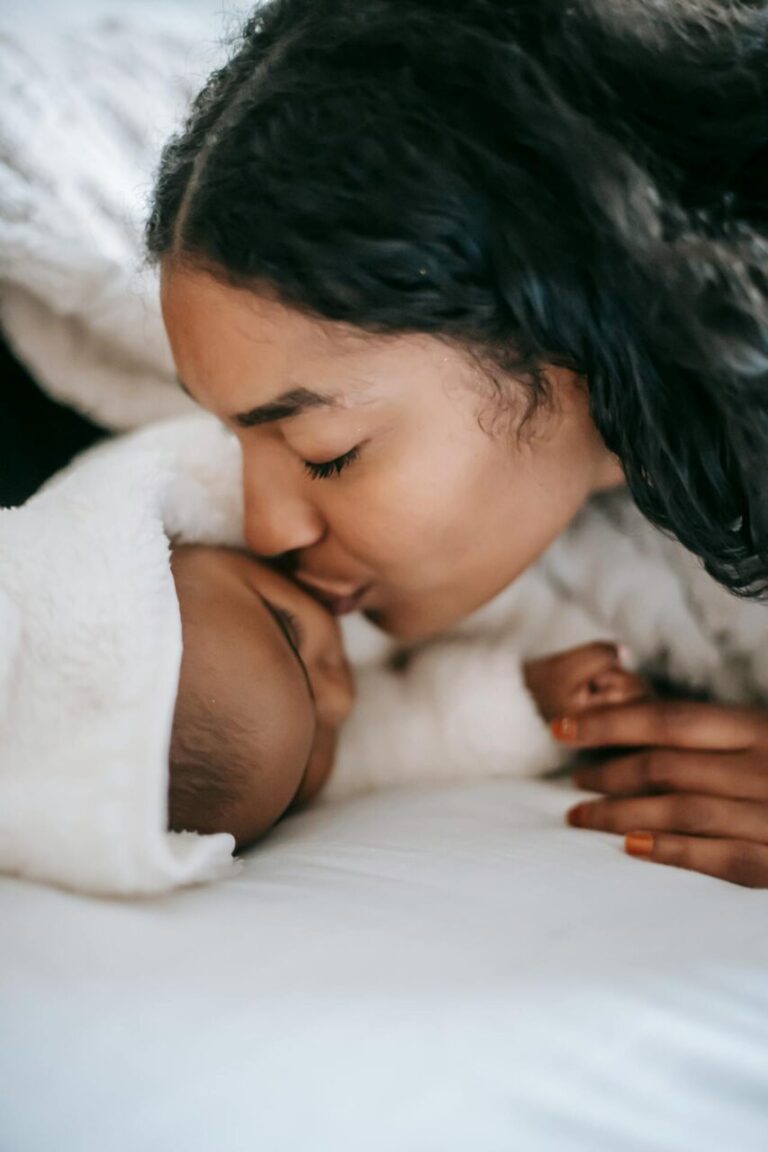 Image of a mother lovingly kisses her swaddled baby on the forehead while lying together on a cozy bed, showing how to be a successful single mom.