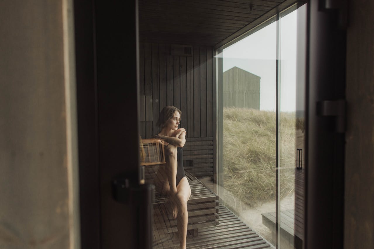 Image of a woman in black swimsuit sitting alone in a wooden sauna looking out a large glass window.
