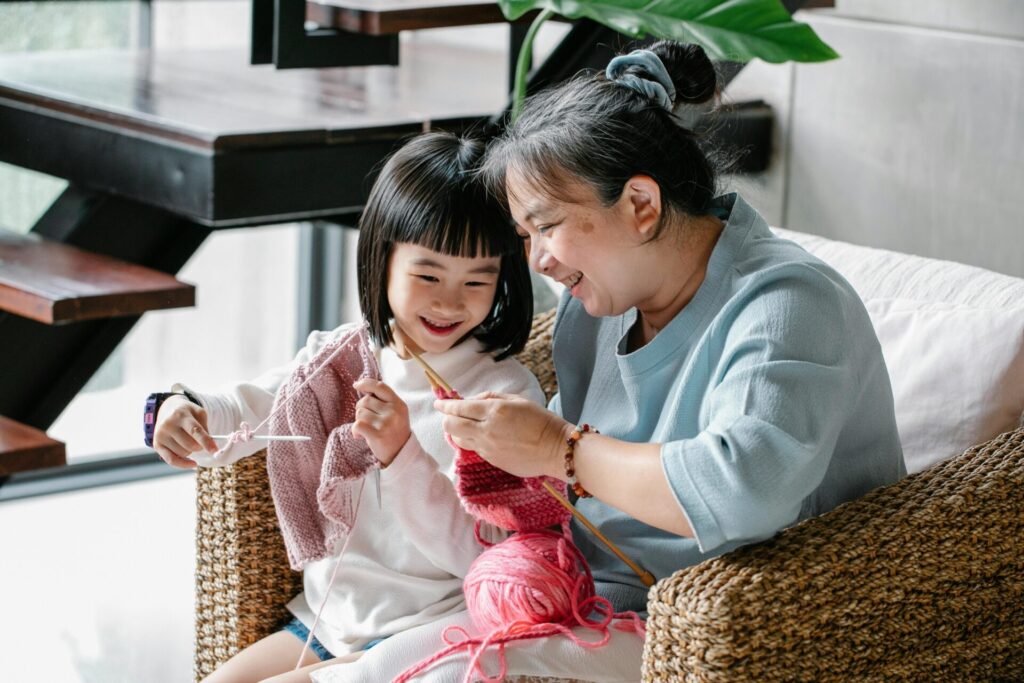 Image of a smiling grandmother teaches her granddaughter knitting with pink yarn at home.