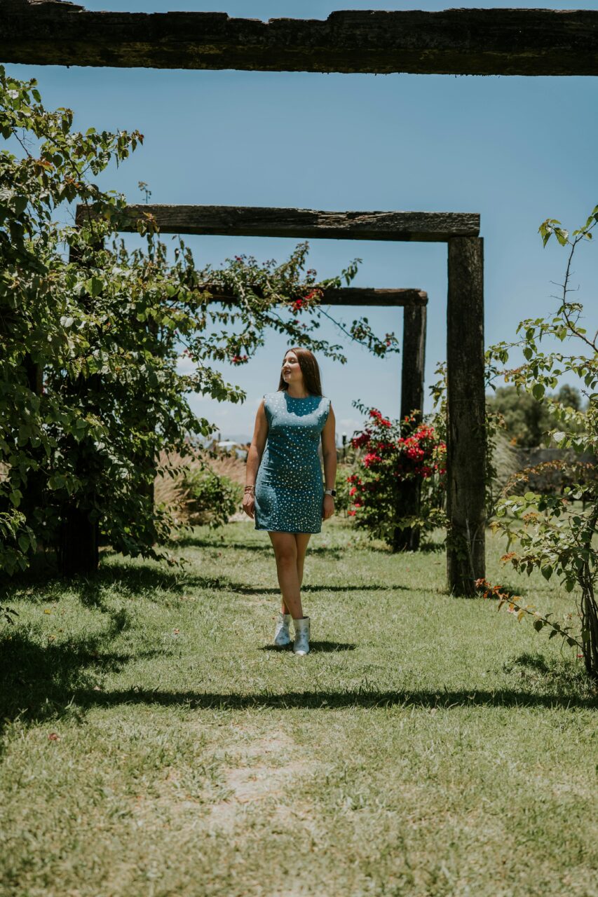 Image of a woman in blue polka dot dress walking in a garden under wooden arches on a sunny day.