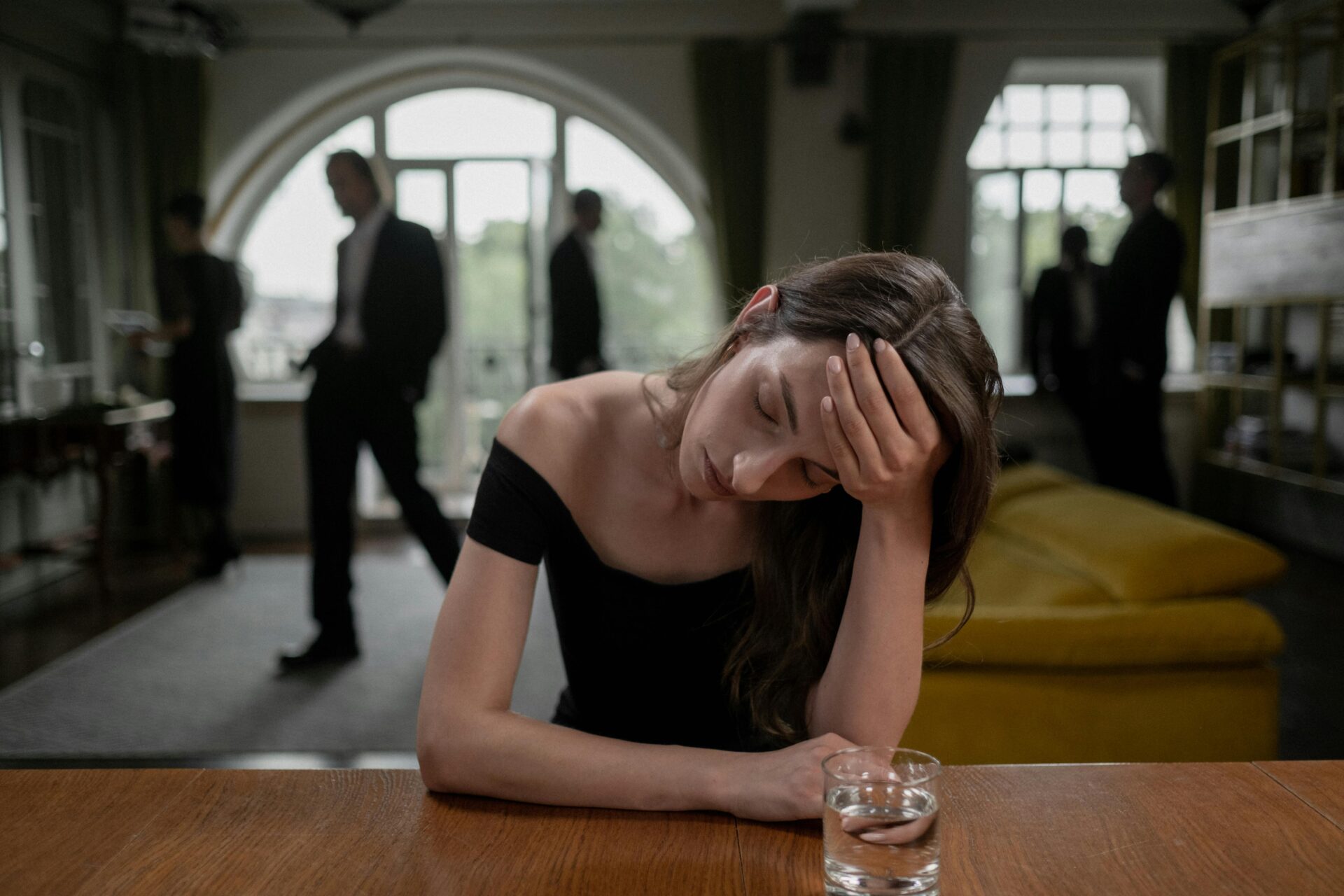 Image of a woman in a black dress sits at a table with her head in hand, looking stressed beside a glass of water, representing I hate networking.