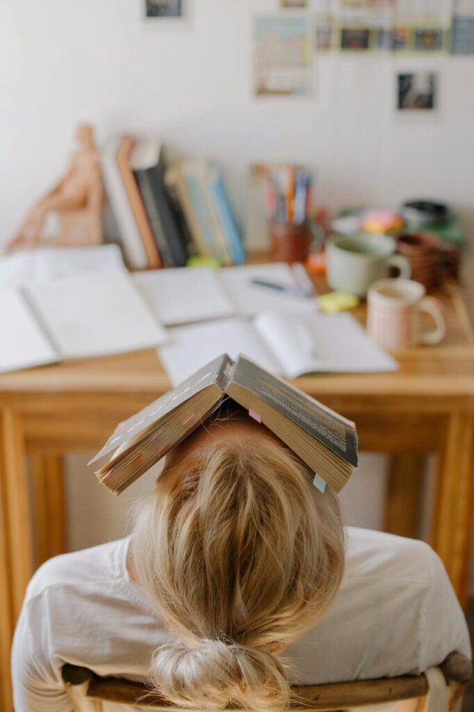 Image of a women resting head with book on face at study desk, showing study stress and exhaustion, showing why do I feel the need To prove myself.