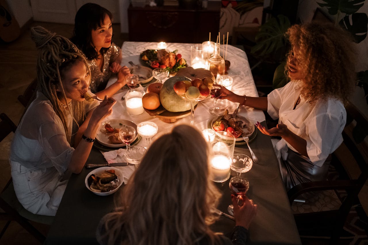 Image of four women enjoying a cozy candlelit dinner together, sharing food, wine, and conversation at a table.