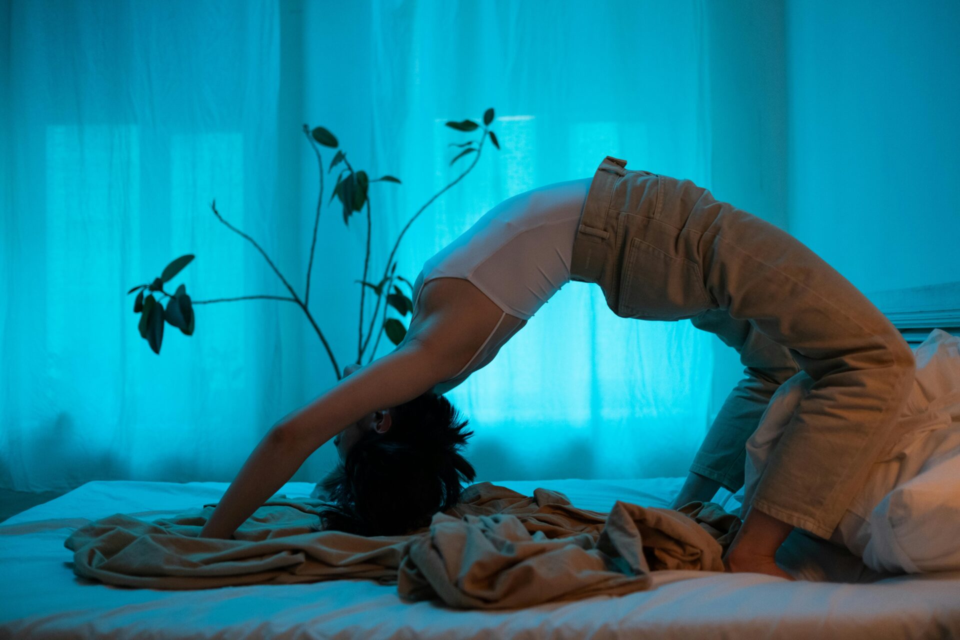 Image of a women doing a backbend yoga pose on a bed with teal curtain background and indoor plant nearby. 
