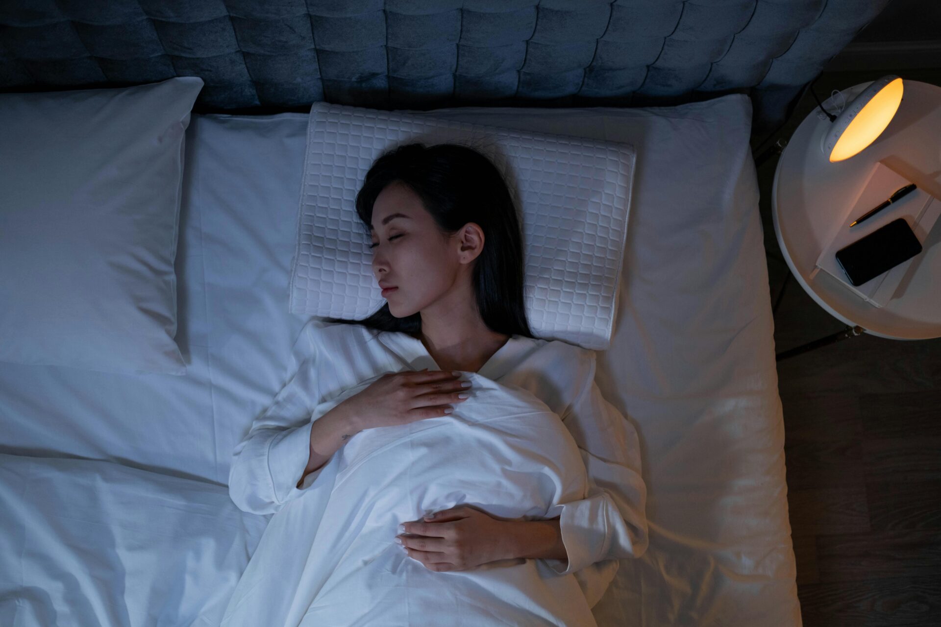 Image of a woman peacefully sleeping in bed at night with a lamp glowing on the bedside table, showing how to glow-up physically.