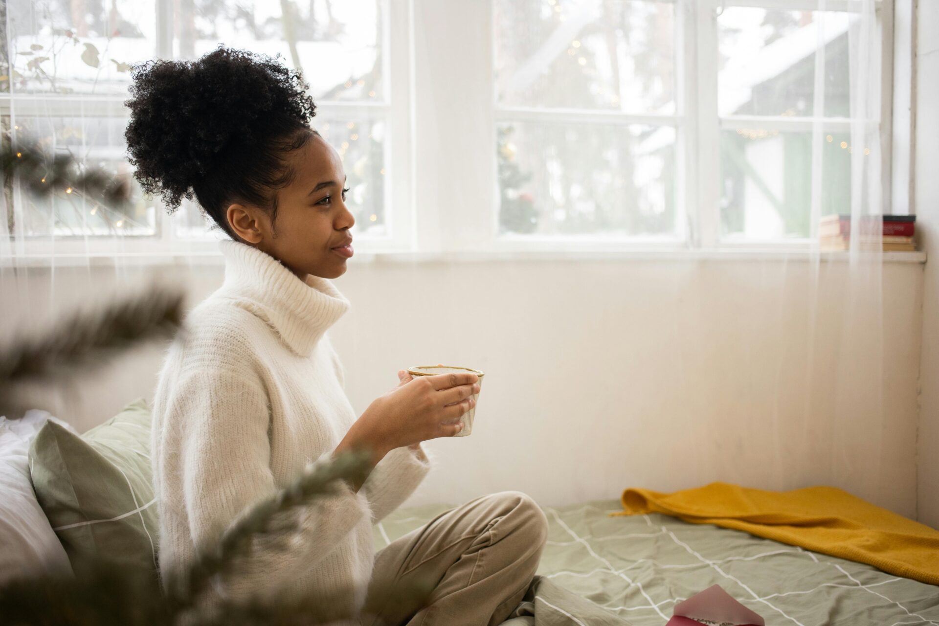 Image of a young woman in white sweater holding a mug, sitting on bed near window with soft light, showing what to wear in South Africa for ladies.