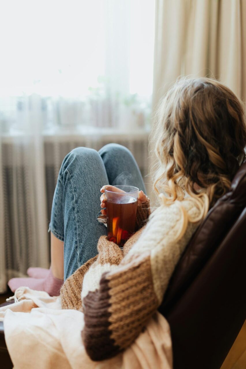 Image of a woman in a cozy sweater relaxes on a chair by the window, holding a warm cup of tea in her hands, representing dissociation vs spacing out.