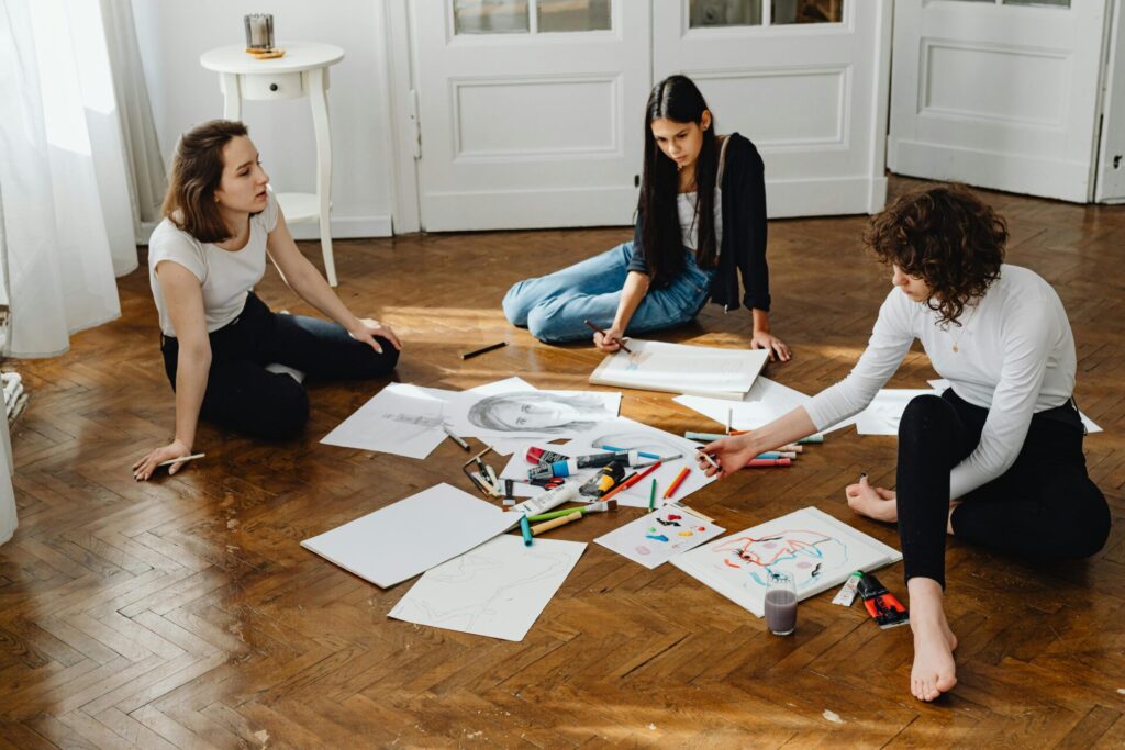 Image of three women sitting on the floor, sketching and painting with art supplies spread around them.