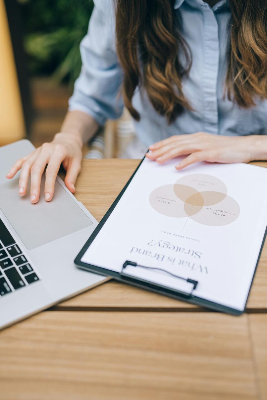 Image of a woman working on laptop with clipboard showing a brand strategy diagram placed on the desk.