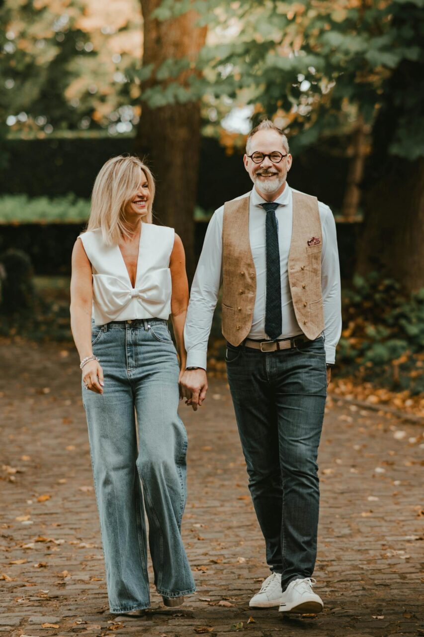 Image of a smiling couple holding hands while walking outdoors on a cobblestone path surrounded by trees.