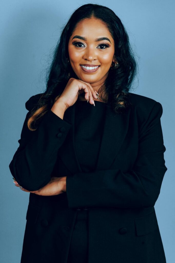Image of a confident woman in a black suit smiling, posing with one hand under her chin against a blue background, showing how can I be independent in a relationship.