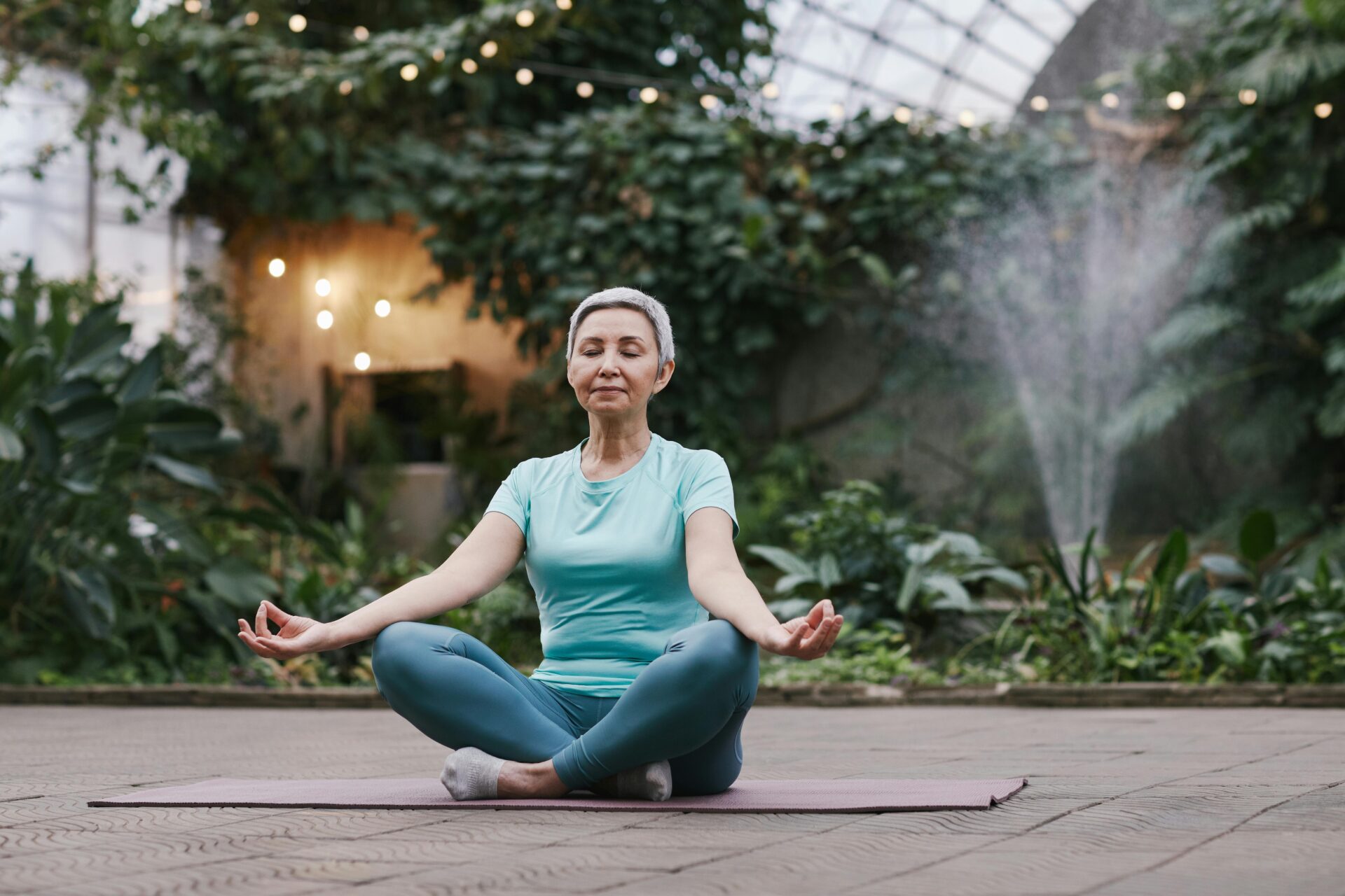 Image of a older woman meditating in lotus pose outdoors on a yoga mat with greenery and fountain in background.