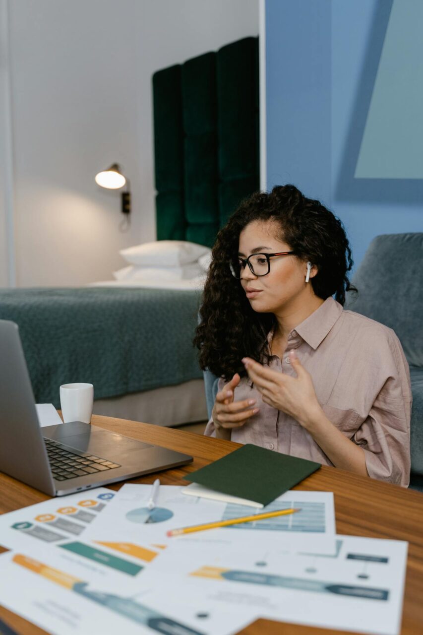 Image of a woman wearing glasses and earphones talking on a video call with charts and laptop on desk.