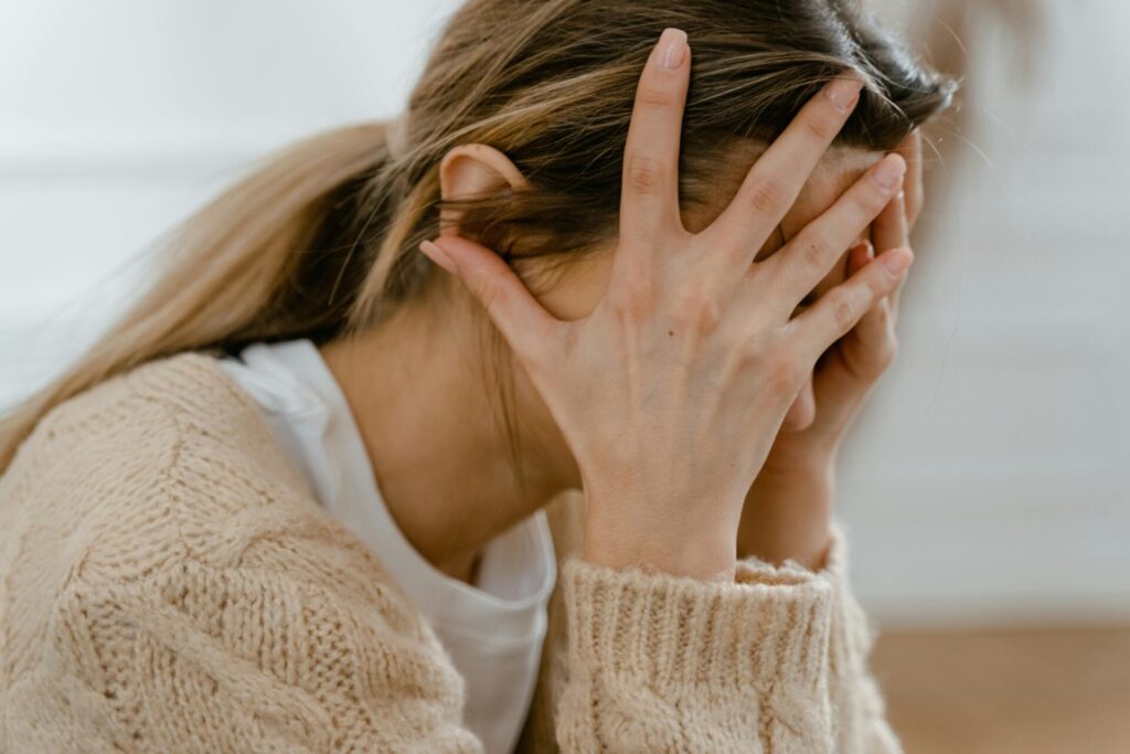 Image of a woman sitting with hands covering face, expressing stress, anxiety, or mental fatigue, showing why do I feel the need To prove myself.