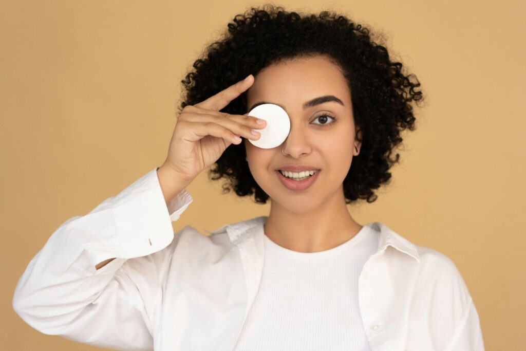 Image of a smiling woman in a white shirt holds a cotton pad over one eye against a beige background.