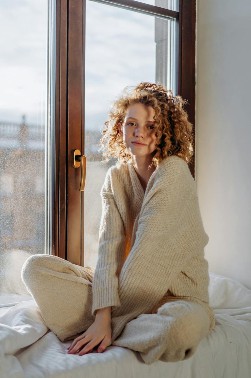 Image of a young woman with curly hair in cozy beige loungewear sits by a sunny window, looking relaxed and calm, showing ADHD zoning out vs dissociation.