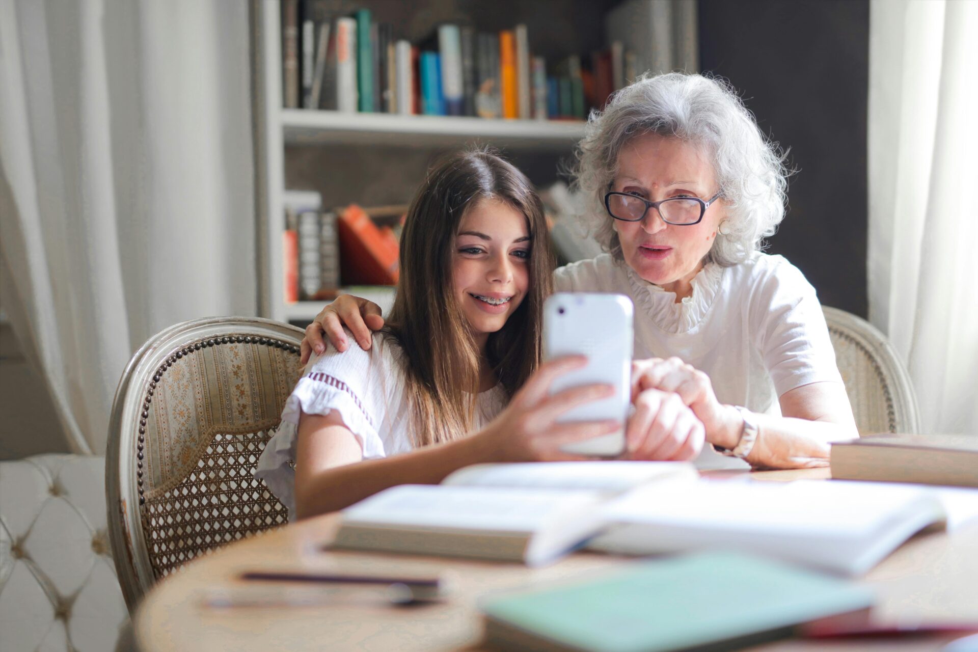 Image of a smiling girl with braces shows her phone to an elderly woman in glasses, sitting together at a desk, showing fun gifts for old people.