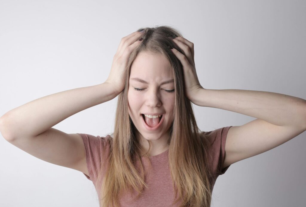 Image of a woman in a pink shirt holds her head with both hands, eyes shut and mouth open, showing frustration.