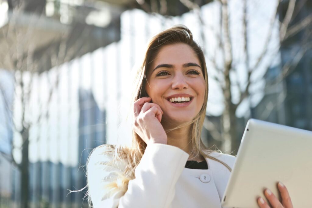 Image of a smiling woman in a white coat holding a tablet and talking outside near modern glass buildings.