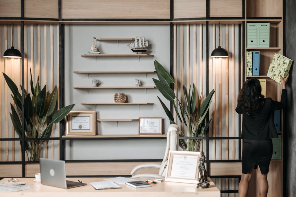 Image of a woman in black outfit placing a file on shelf in modern office with laptop, plants, and decor.
