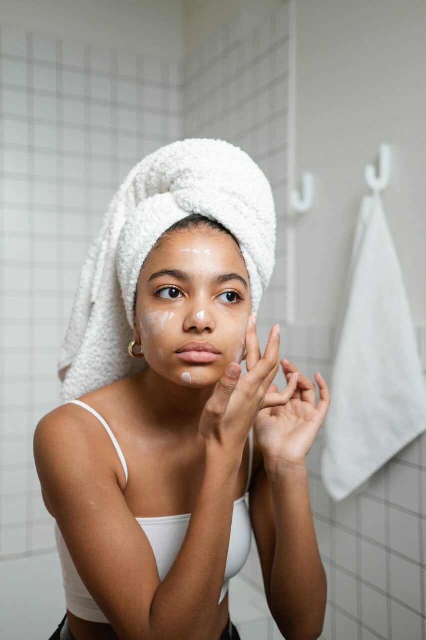 Image of a woman with towel on head applying skincare cream to her face in a bathroom mirror, representing glow-up checklist for women.