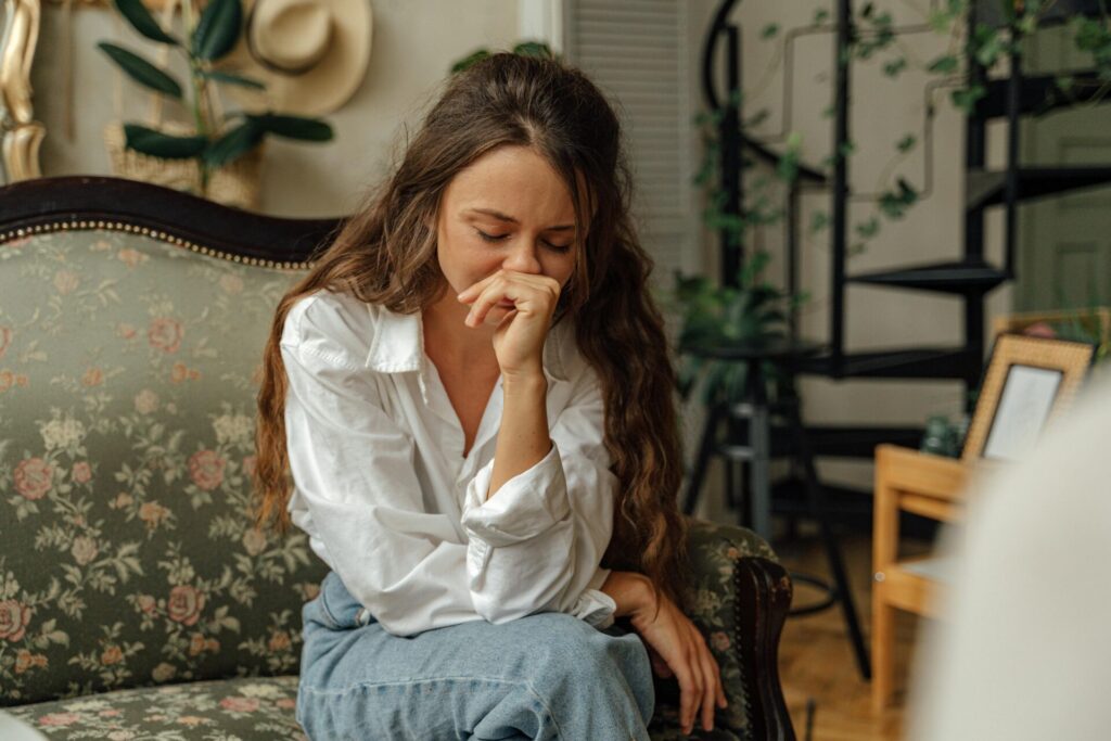 Image of a young woman in a white shirt sits on a floral sofa, leaning forward with her hand near her face, worried.
