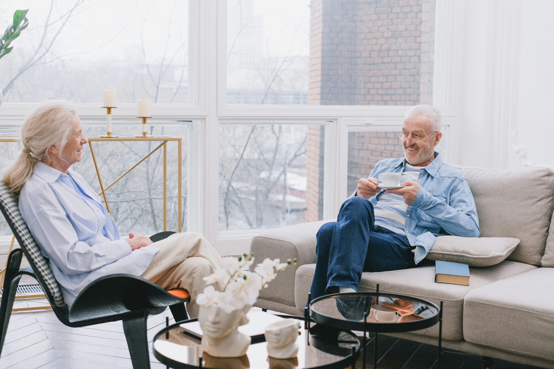 Image of a senior couple sitting in a bright living room, smiling and enjoying tea together, showing presents for old people