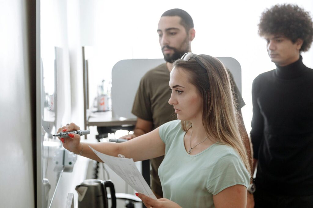 Image of a young woman writing on whiteboard with marker while two men watch attentively in a modern workspace, showing how to find your purpose.