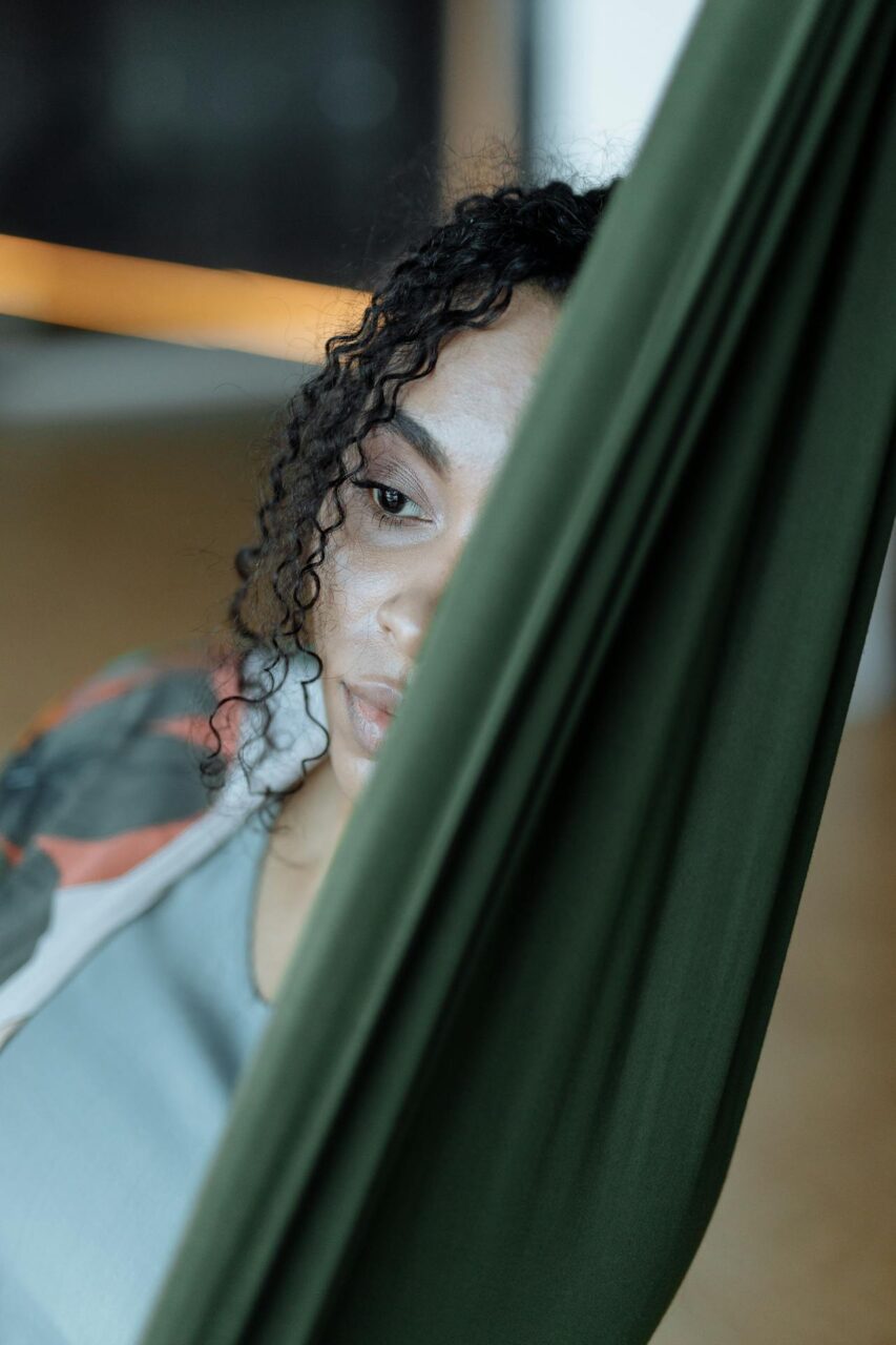 Close-up image of a thoughtful woman gazes past green aerial yoga fabric, her face partly hidden, creating a serene mood, showing zoning out vs dissociation.