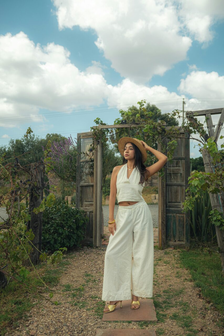 Image of a woman in white outfit and straw hat poses gracefully outdoors near rustic wooden doors under blue sky.