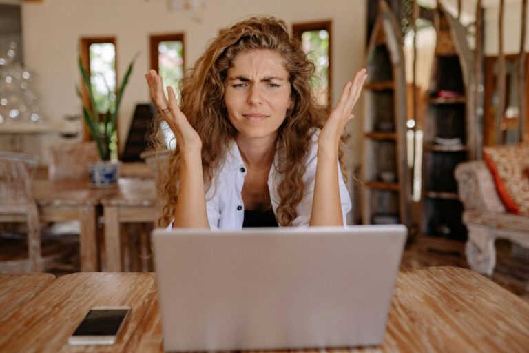 Image of a frustrated woman with curly hair sits at a laptop, raising hands in confusion at a wooden table, showing I hate networking.