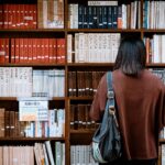 Image of a woman with a bag stands in front of a library bookshelf filled with neatly arranged books, showing 12 self-help books for moms recommended in 2026.