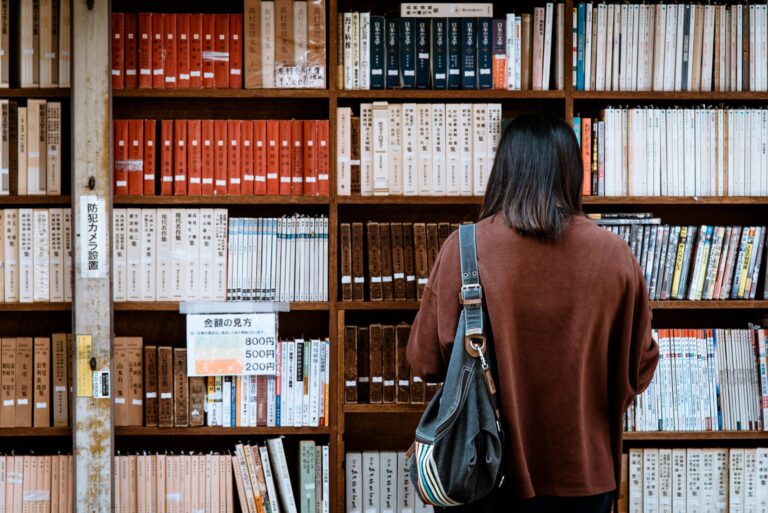 Image of a woman with a bag stands in front of a library bookshelf filled with neatly arranged books, showing 12 self-help books for moms recommended in 2026.