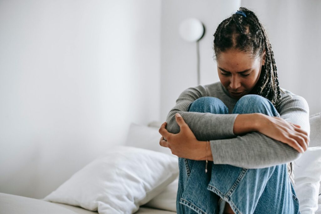 Image of a woman sitting on bed hugging knees, looking sad and stressed in a quiet white room, showing mental load in a relationship.