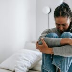 Image of a woman sitting on bed hugging knees, looking sad and stressed in a quiet white room, showing mental load in a relationship.
