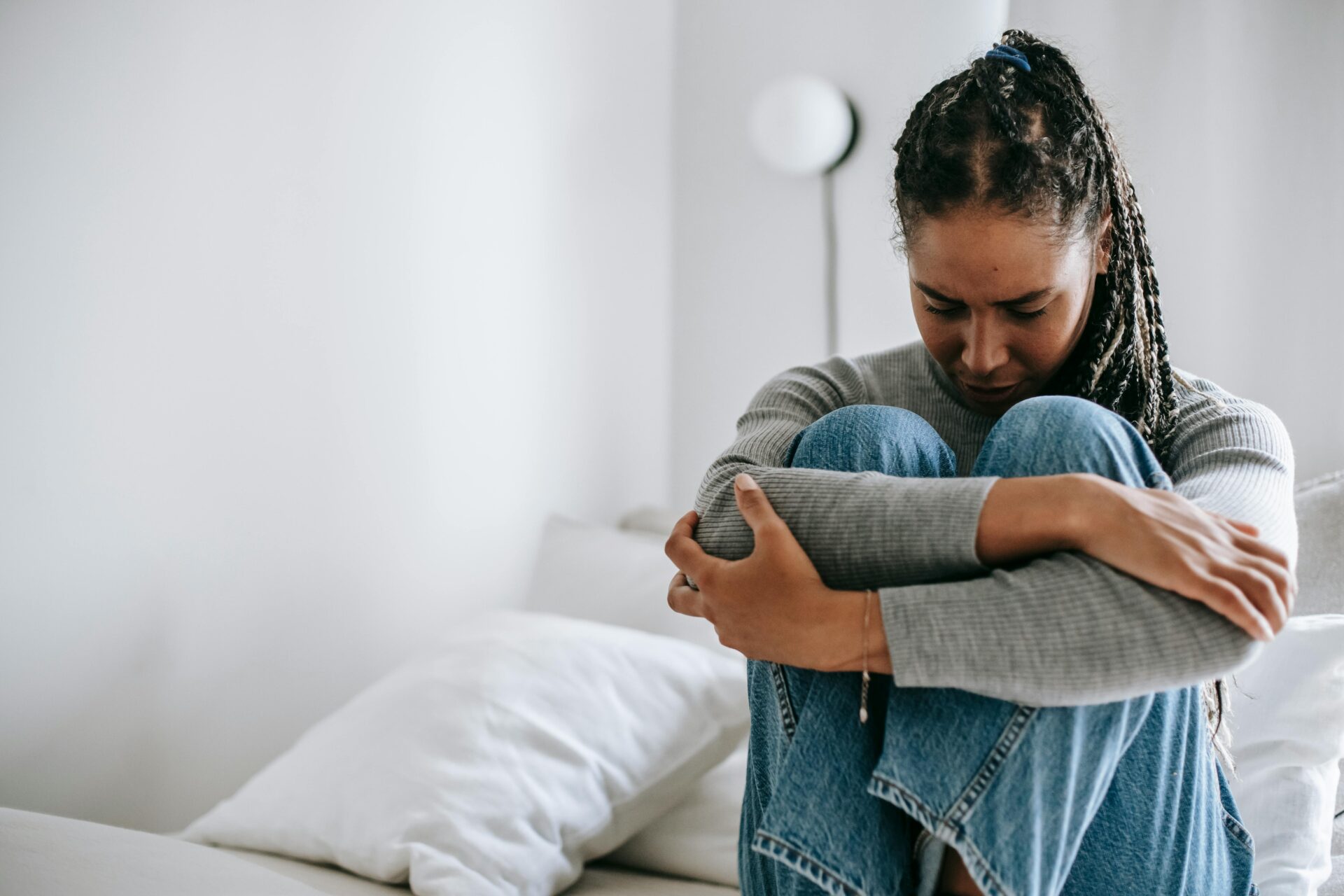 Image of a woman sitting on bed hugging knees, looking sad and stressed in a quiet white room, showing mental load in a relationship.