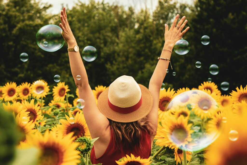 Image of a woman in a sunflower field raising arms joyfully with bubbles floating around her.