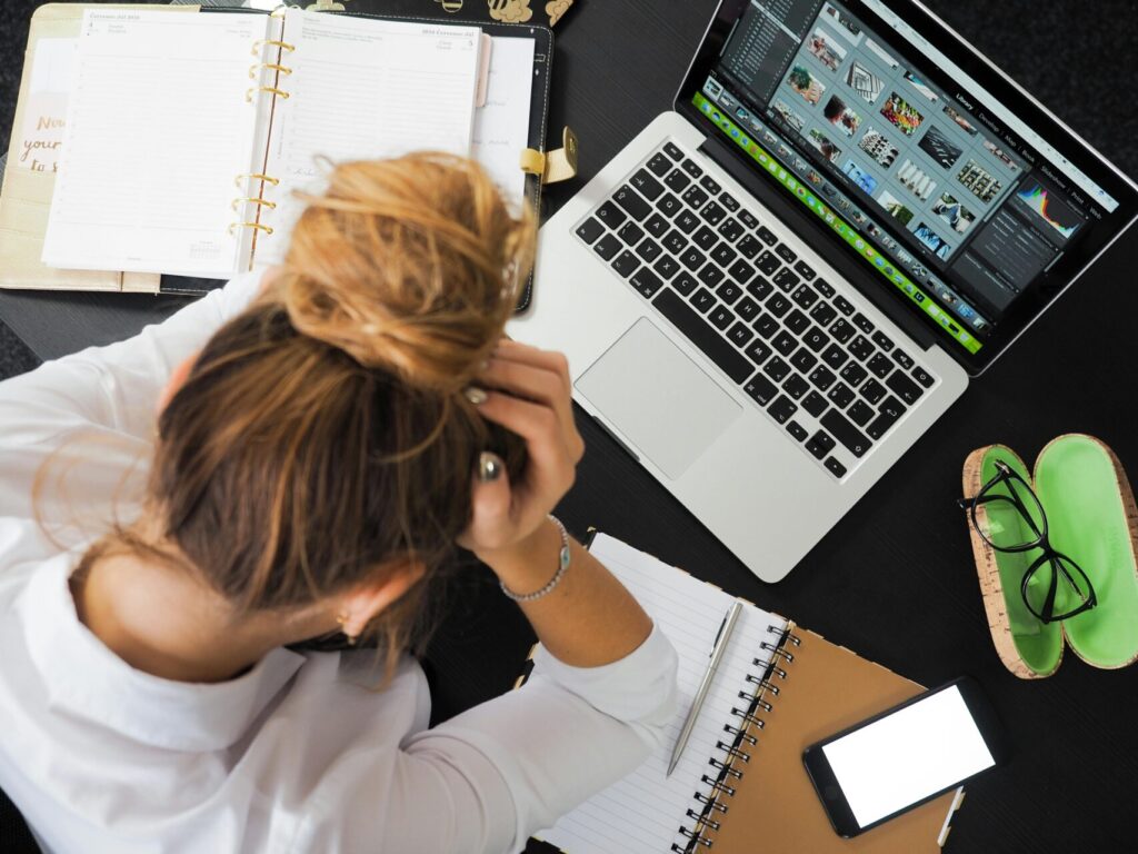 Image of a woman stressed at desk with laptop, notebook, planner, and phone, holding her head in hands, showing 30-Day stress relief journal prompts.