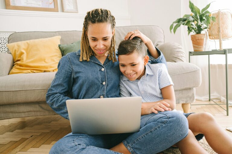Image of a mother and son sitting on floor, smiling while looking at laptop together in cozy living room, showing madewell.
