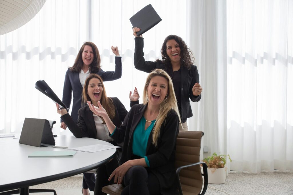 Image of four women in business attire celebrating a success, smiling and cheering with documents in hand, showing how to find Huntsville support groups.
