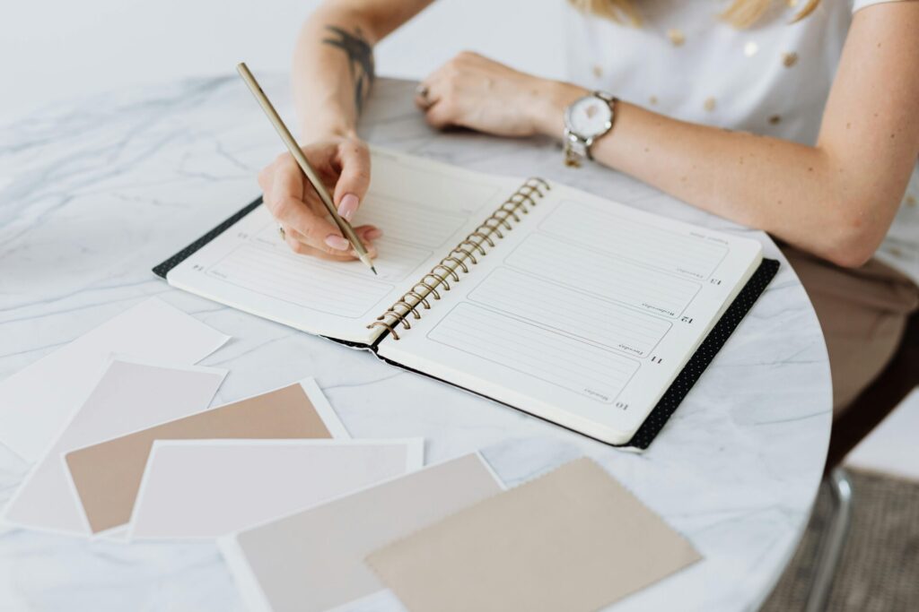 Image of a women writing in a spiral planner on a marble table with color swatches spread around, showing 30-Day stress relief journal prompts.