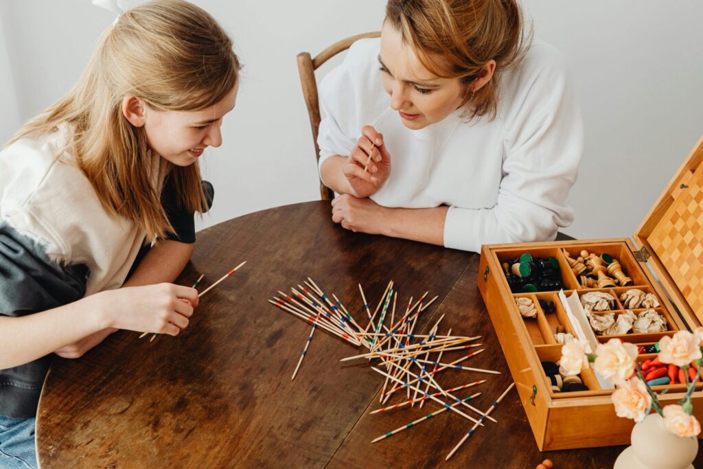 Image of two women's smiling while playing pick-up sticks at a wooden table with a box of board game pieces.