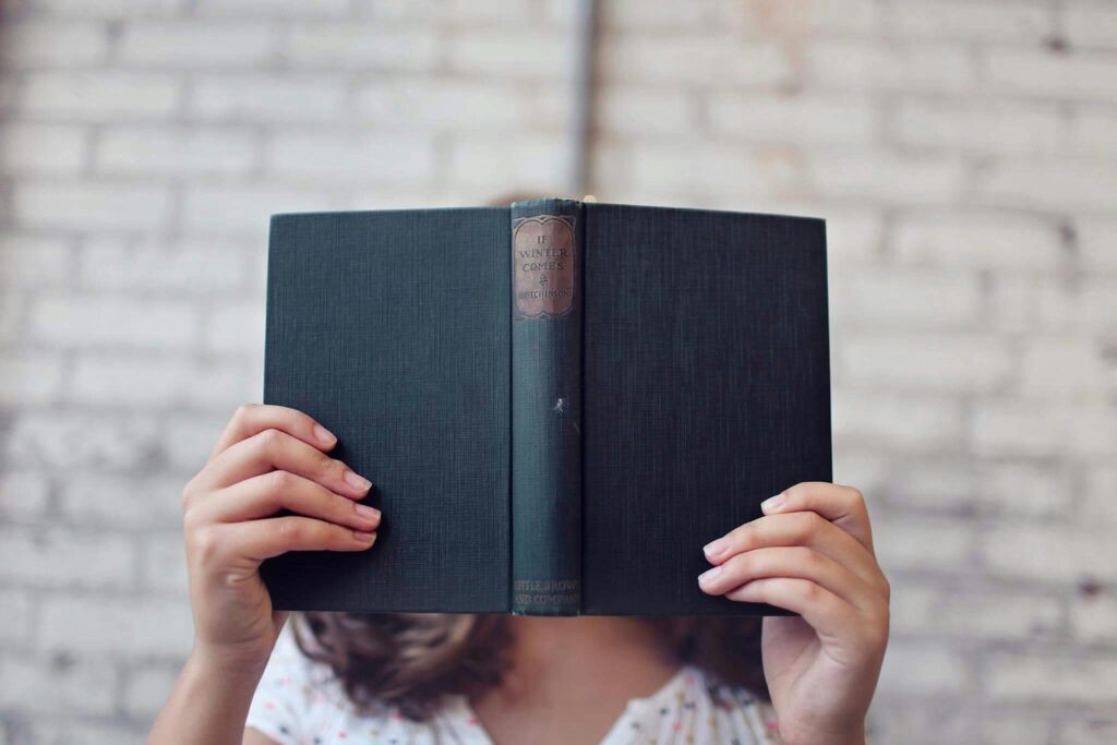 Close-up image of a women holding a book with a black cover, wearing white top in white brick background.