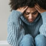 Image of a woman in blue sweater holding head in distress, sitting with eyes closed, showing how to handle seasonal depression in Huntsville winters.